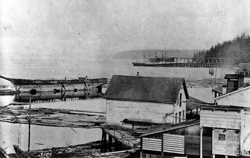 Black and white image showing a wooden ship without masts in the water next to waterfront buildings.