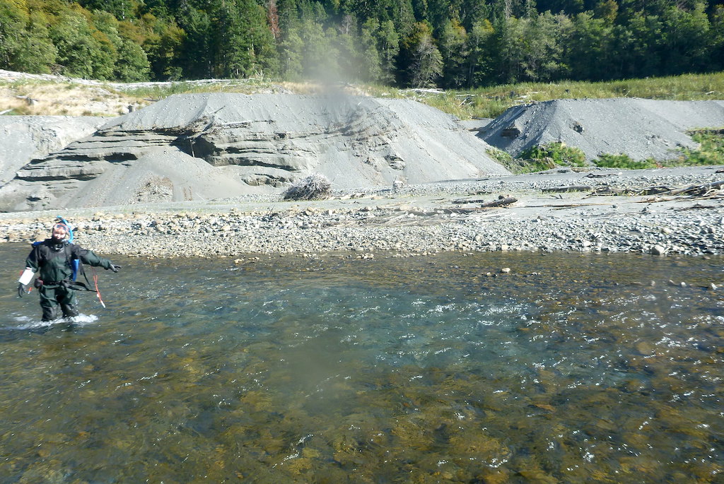 A person wearing snorkeling gear standing in a river pointing to an area with lighter colored river rocks.
