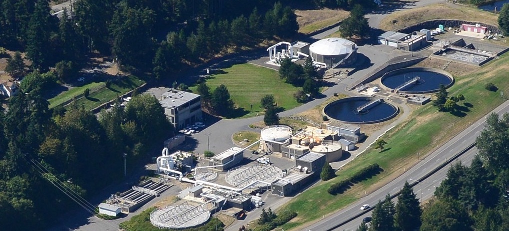 Aerial view of a wastewater treatment plant with circular and rectangular tanks, surrounded by trees and a road on the right.