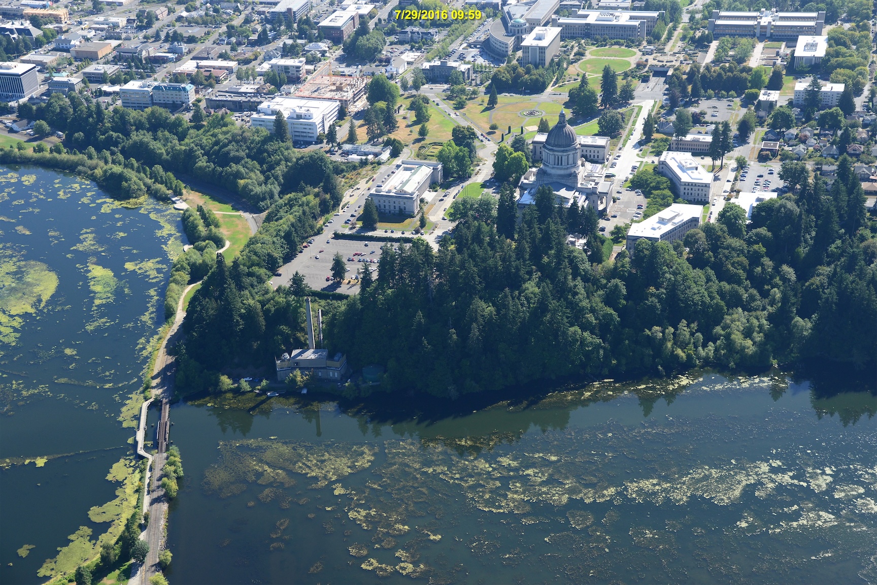 Aerial view of a city with a domed building, bridge on the left, and water with algae patches in the foreground. Dated 7/29/2016.