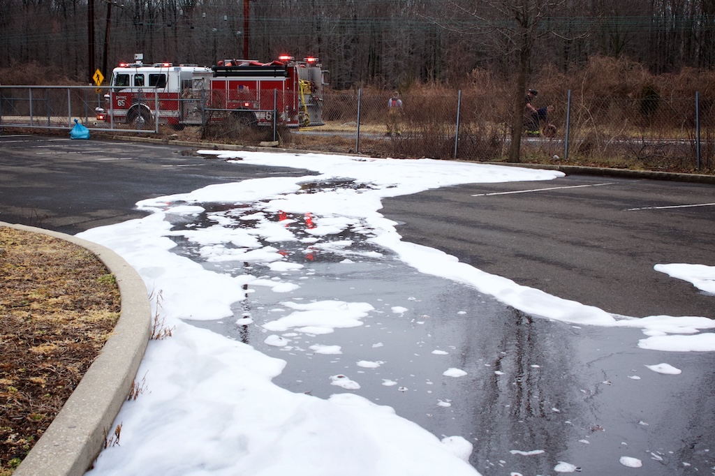 Fire truck in the background with white firefighting foam covering pavement and pooling on surface in the foreground.