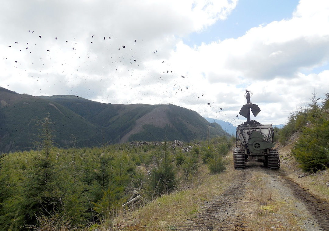 Truck dispersing biosolids onto young forest trees forest from a road with mountains in background.