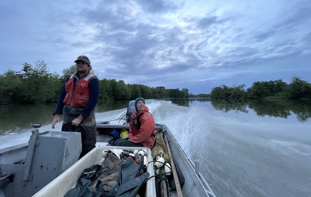 Two people in an open motor boat travel on a calm river.