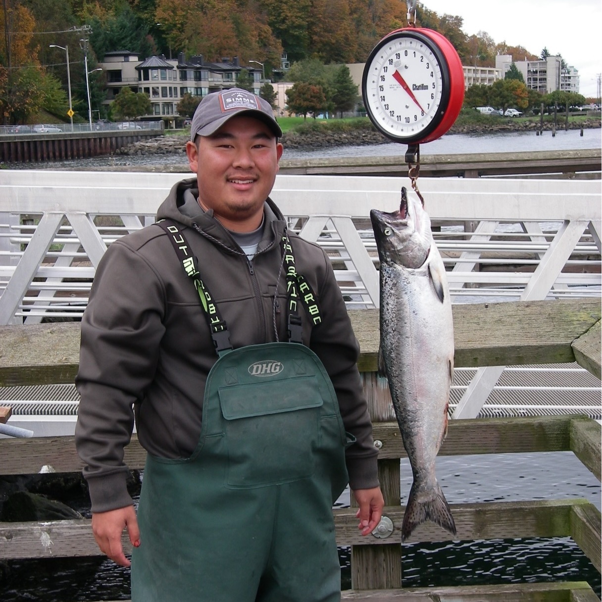 A person in waders standing next to a fish hanging from on a scale to weigh it.