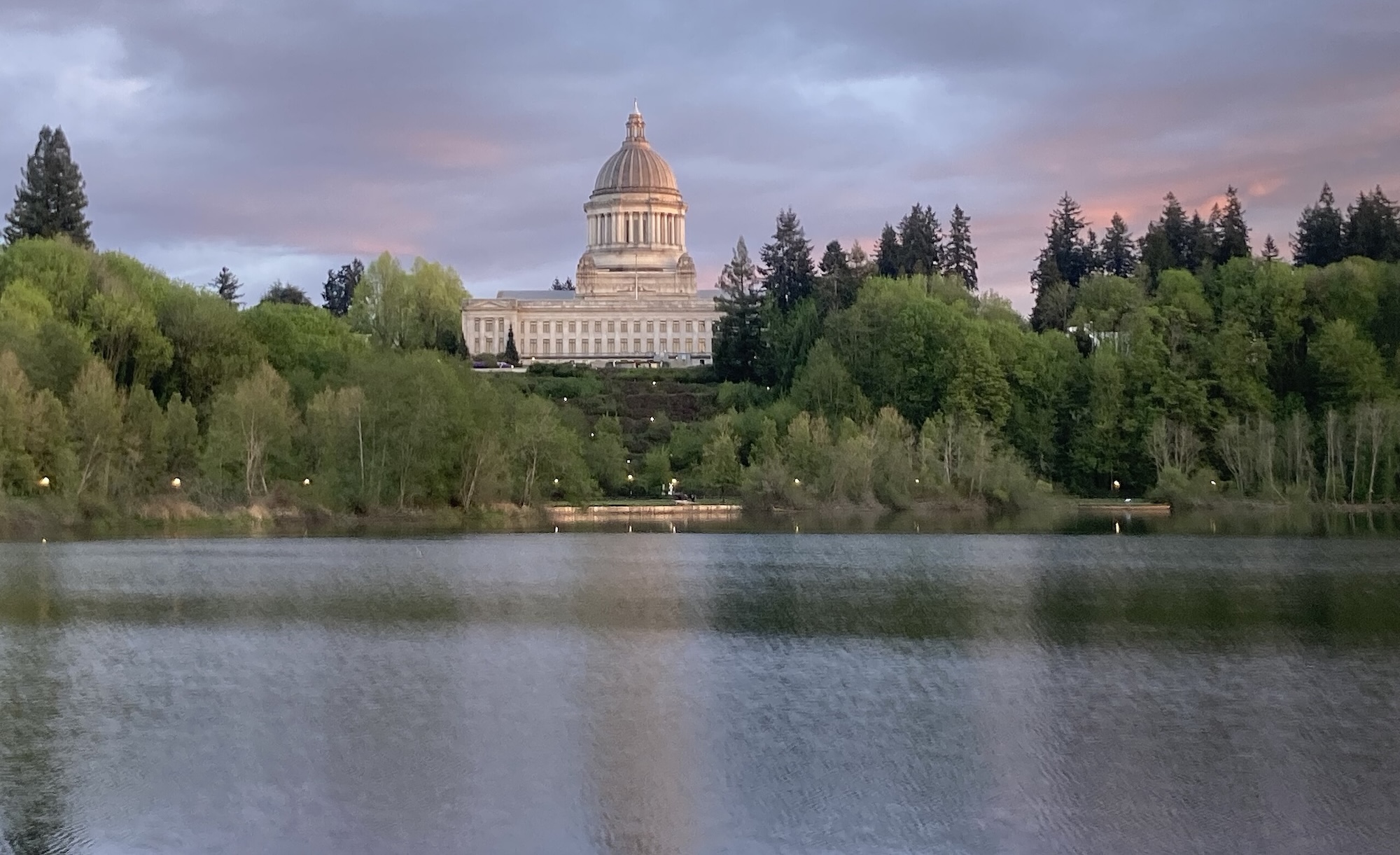 Washington State Capitol in Olympia viewed across Capitol Lake, with dome framed by trees under a pink and purple sunrise or sunset sky.
