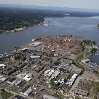 Aerial view of Budd Inlet showing a wastewater treatment plant with circular clarifiers, industrial waterfront facilities, marinas, and forested shoreline with mountains in the background.