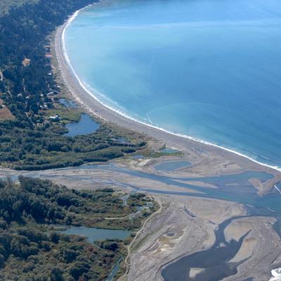 Aerial view of curved coastline showing forested hills, winding river delta with mudflats, and long sandy beach meeting bright blue ocean waters.