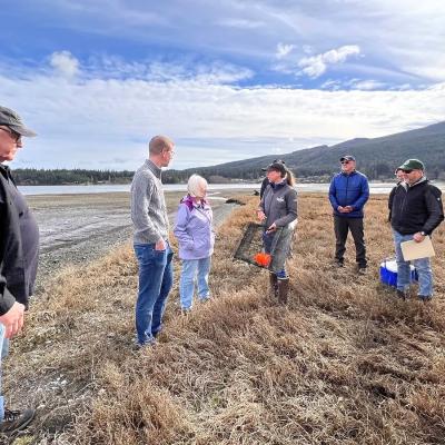 Group of seven people gathered on a tidal flat or salt marsh area examining crab traps, with mountains and water visible in the background under a partly cloudy sky.