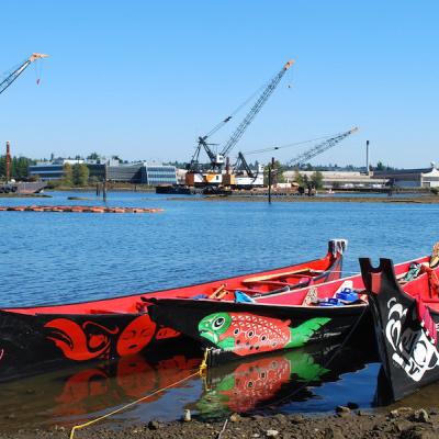 Brightly painted canoes on river shore with construction cranes in background.