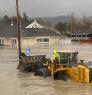 Two people operating a bulldozer at the intersection of two flooded streets in Sumas, Washington. In the background, partially submerged cars are parked in front of the library.
