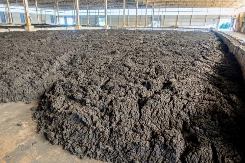 A large indoor facility containing a wide expanse of dark, thick, biosolids spread across the floor beneath a roof structure.