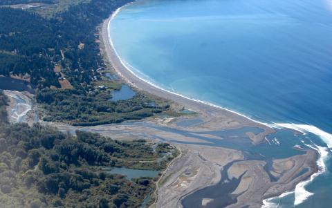 Aerial view of curved coastline showing forested hills, winding river delta with mudflats, and long sandy beach meeting bright blue ocean waters.