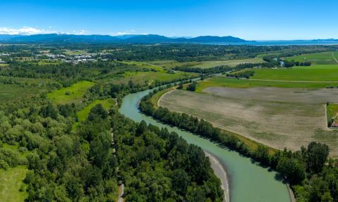 Aerial view of a river flowing through farmland buffered on both sides by trees and other vegetation, with islands and Puget Sound in the distance.