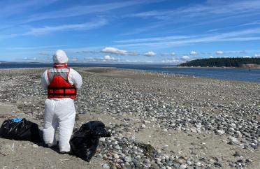 A person wearing a Tyvek suie and orange vest standing on a beach next to two full garbage bags. Water, land, and blue sky in the distance