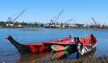 Brightly painted canoes on river shore with construction cranes in background.