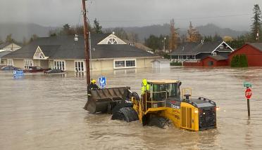 Two people operating a bulldozer at the intersection of two flooded streets in Sumas, Washington. In the background, partially submerged cars are parked in front of the library.