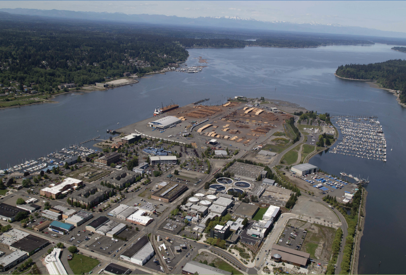 Aerial view of Budd Inlet showing a wastewater treatment plant with circular clarifiers, industrial waterfront facilities, marinas, and forested shoreline with mountains in the background.