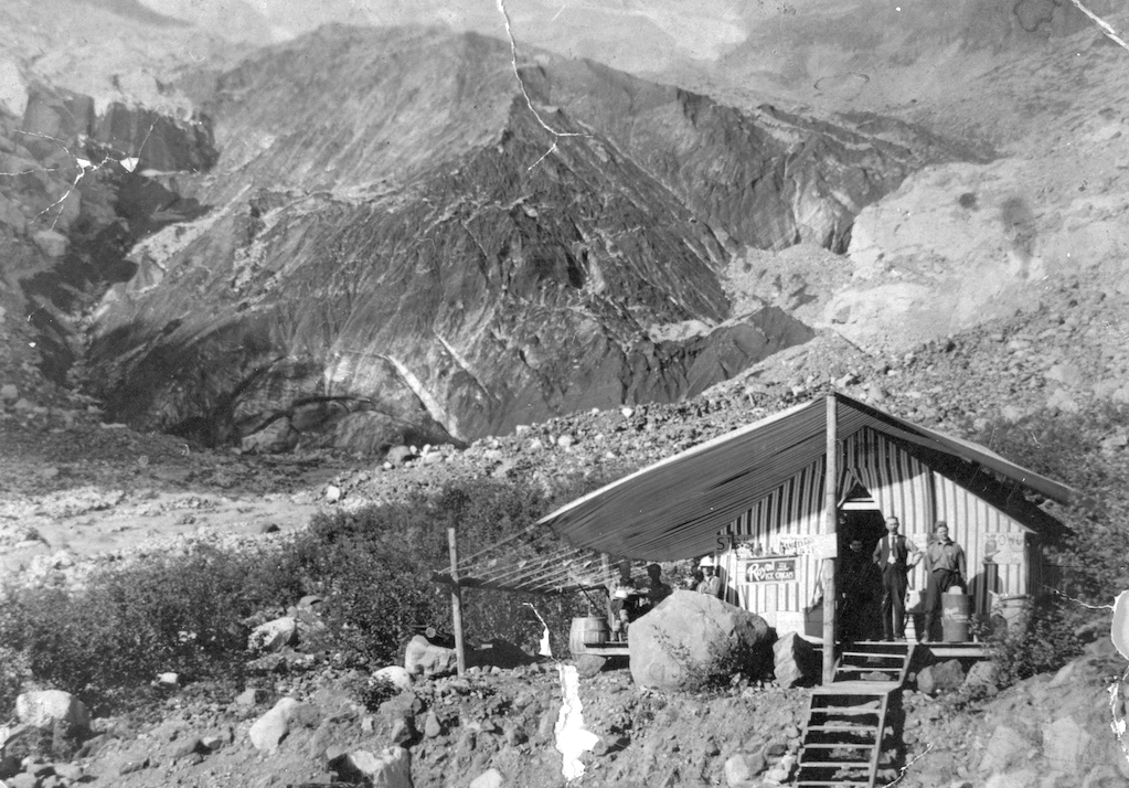 Black and white photo of two men standing in front of a canvas shack with mountain and glacier behind it.