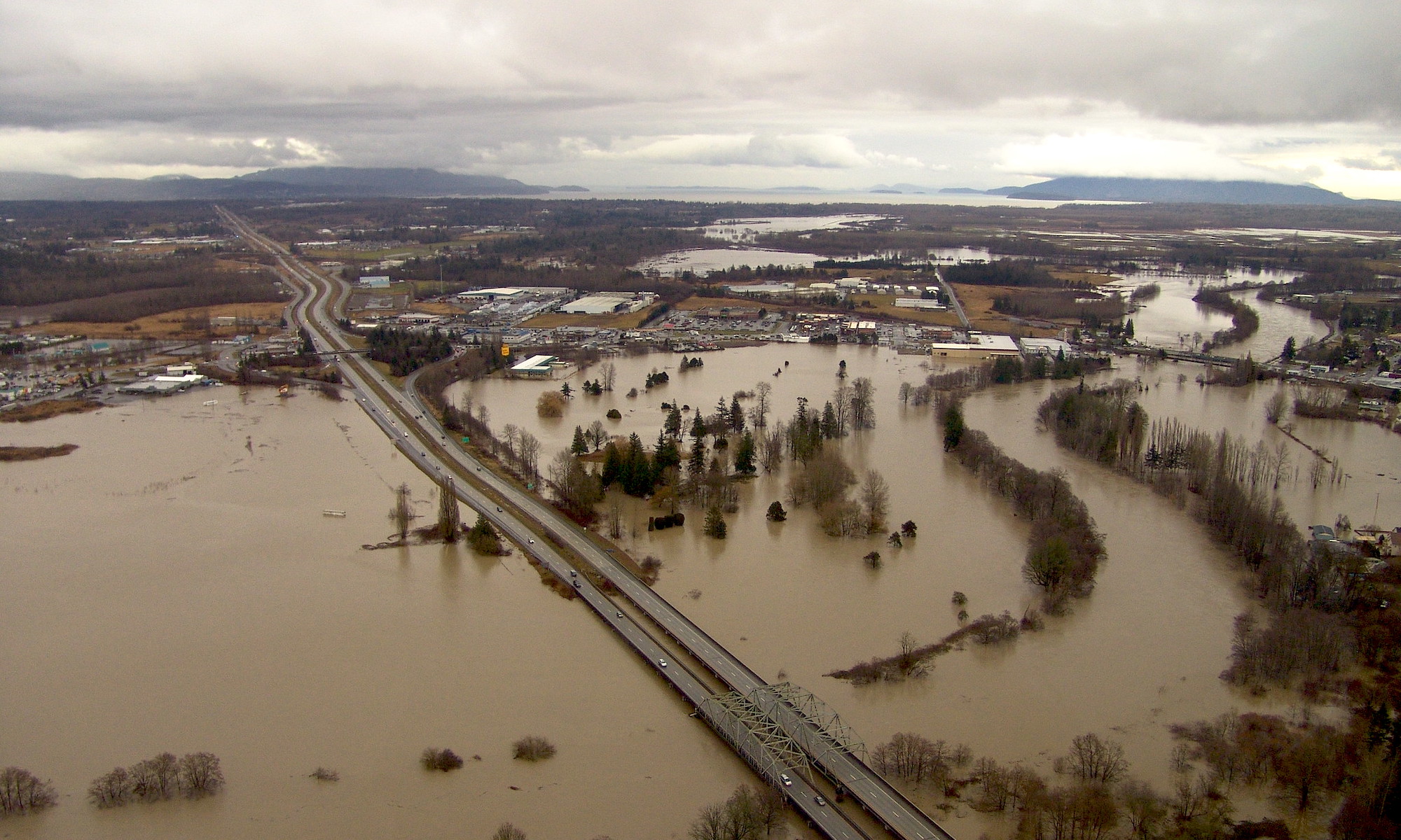 							Aerial view of Interstate 5 stretching across a large area of land covered by brown flood waters from the Nooksack River in the foreground with mountains and Puget Sound in the distance and grey skies above.							