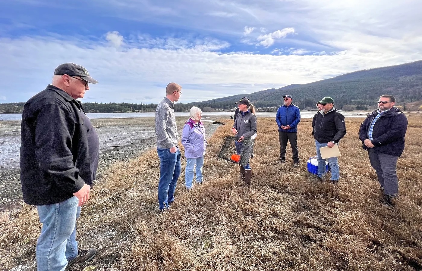 Group of seven people gathered on a tidal flat or salt marsh area examining crab traps, with mountains and water visible in the background under a partly cloudy sky.